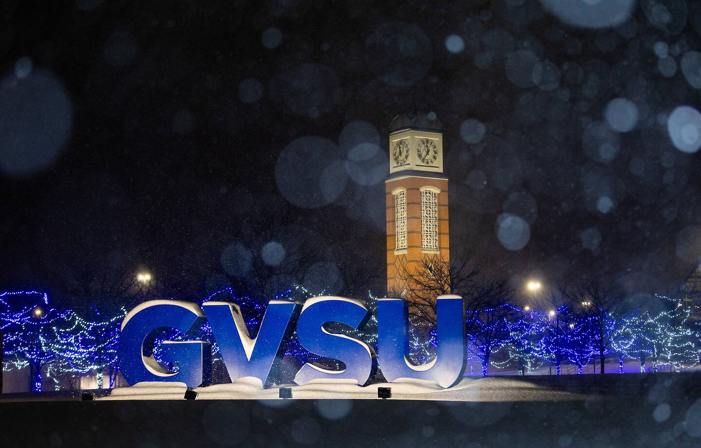 At night, illuminated blue "GVSU" letters stand in front of a clock tower, surrounded by snow and festive blue lights on trees.